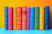 Row of colorful hardcover books standing on turquoise surface against yellow wall background, arranged in rainbow spectrum order creating vibrant composition.