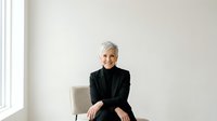 Senior woman with short gray hair, wearing black blazer and black turtleneck, sitting on beige chair against white wall. Professional portrait for business, corporate, and lifestyle use.