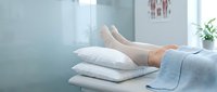 Woman resting legs on stack of white pillows in bright bedroom, wearing light blue blanket, relaxation and comfort concept for wellness and home lifestyle.