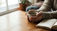 Person holding warm beverage while reading book at wooden table by window, cozy indoor relaxation scene for lifestyle and wellness content.