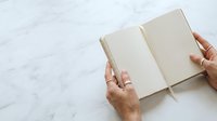 Person with medium skin tone holding open blank notebook on marble surface for writing, journaling, planning, or creative projects.