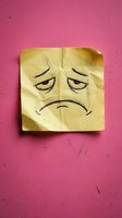 Crumpled yellow sticky note with hand-drawn sad face expression on textured pink background representing depression, disappointment and negative emotions.