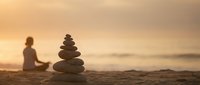 Zen stone stack on beach sand with blurred woman meditating at golden sunset, creating peaceful mindfulness scene for wellness and spiritual balance concepts.