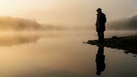 Silhouette of contemplative man standing by misty lake at golden sunrise with perfect reflection in calm water creating peaceful morning atmosphere.