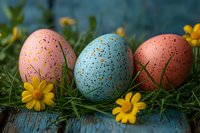 Speckled pastel Easter eggs in pink and blue decorated with gold details, arranged on green grass with yellow daisies against rustic wooden background.