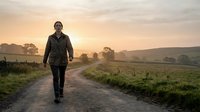 Young woman with dark hair and medium complexion wearing black jacket walking on rural dirt road at sunrise through countryside landscape with rolling hills and morning mist.