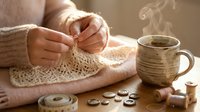 Woman hands knitting cream wool sweater with coffee cup and buttons on wooden table. Cozy handmade crafting hobby scene for relaxation and creativity projects.
