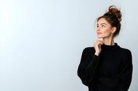 Young woman in black sweater with messy bun hairstyle looking thoughtfully to side against light background, hand on chin in contemplative pose.