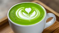 Green matcha latte with white heart-shaped latte art foam design in ceramic cup on wooden surface, close up view showing vibrant color and detailed pattern.