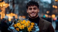 Young Hispanic man in brown blazer and black turtleneck holding bright yellow daffodil bouquet, smiling warmly against blurred evening city lights and bokeh background.