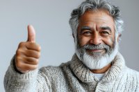 Cheerful mature man with gray beard and hair wearing knit sweater showing thumbs up gesture against light background, expressing positivity and approval.