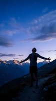 Male hiker with open arms celebrating freedom on mountain trail under starry twilight sky with dramatic clouds and alpine peaks in background.