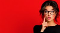 Young Asian woman with glasses and surprised expression against vibrant red background, wearing black top, finger near lips in thoughtful gesture. Messy bun hairstyle.