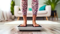 Woman's bare feet standing on weight scale wearing patterned leggings. Modern living room interior with sofa and plants in background. Healthy lifestyle concept.