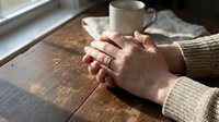 Clasped hands of a woman wearing a gold ring resting on a rustic wooden table near a window, with a ceramic mug in soft background light.