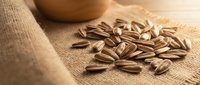 Roasted sunflower seeds scattered on natural burlap fabric with wooden bowl, suitable for food photography, healthy snacking, nutrition content, and culinary design projects.