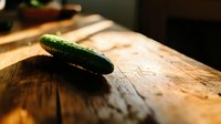 Fresh green cucumber on rustic wooden cutting board in natural sunlight, creating dramatic shadows and warm atmosphere for food photography.