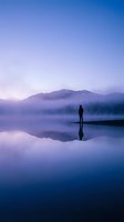 Solitary figure standing on misty lake shore at dawn with mountain silhouettes and purple sky reflection creating peaceful meditation scene.