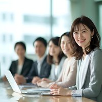 Smiling Asian businesswoman leading team meeting in modern office conference room with diverse colleagues in background.