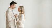 Young couple in beige casual wear holding hands and looking at each other with tender expressions against neutral background for lifestyle content.