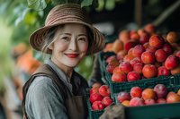 Asian female farmer in straw hat and apron smiling while standing next to fresh organic nectarines and peaches at local farmers market produce stand.