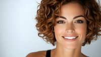 Young Caucasian woman with curly brown hair smiling warmly at camera against neutral background. Professional headshot for business marketing materials.