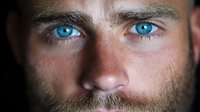 Close up portrait of young Caucasian male with intense bright blue eyes and short beard against dark background, dramatic lighting emphasizes facial features.