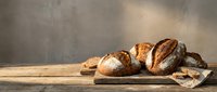 Fresh artisan bread loaves with crusty golden brown exteriors arranged on rustic wooden table against textured concrete wall background for bakery display.