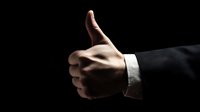 Male hand in formal business suit giving thumbs up gesture against dark background, symbolizing approval, success, and positive feedback.