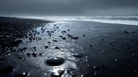 Moody coastal landscape with dark volcanic sand beach, wet surface reflecting overcast sky, scattered rocks and pebbles along shoreline during stormy weather conditions.