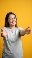 Young woman with long dark hair and fair complexion wearing a light gray t-shirt, smiling warmly with arms outstretched in a welcoming gesture against a bright yellow background.