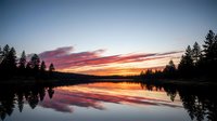 Serene lake at sunset with vibrant orange and pink sky reflecting on calm water, surrounded by evergreen forest silhouettes, peaceful nature landscape.