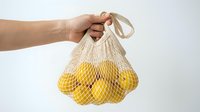 Hand holding a reusable cotton mesh bag full of fresh yellow lemons on a light background, eco-friendly shopping and zero waste concept.