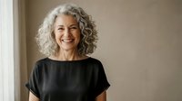 Happy mature woman with curly gray hair smiling confidently in black top against neutral background for lifestyle and wellness content.