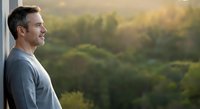 Middle-aged man with dark hair and facial stubble gazing thoughtfully out window at forested landscape during golden hour, wearing casual gray shirt, contemplative mood for lifestyle and wellness content.