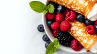 Fresh berries and thin rolled crepes garnished with mint leaves on white plate, closeup view of sweet breakfast or dessert serving arrangement.