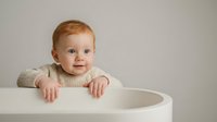 Adorable redhead baby with blue eyes sitting in white high chair wearing beige knit sweater against neutral background for family lifestyle content.