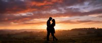 Silhouetted couple kissing in open field during dramatic golden sunset with sparkling light effects and mountains in background.