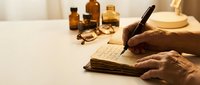 Hands writing with vintage fountain pen in old notebook surrounded by antique glass bottles and brass compass on wooden desk for historical storytelling.