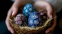 Decorated Easter eggs with traditional folk patterns in blue and brown colors resting in straw nest held by female hands. Dark moody background creates festive atmosphere.