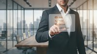 Middle-aged male professional in business suit holding transparent digital interface displaying data analytics metrics in modern office with city skyline view.