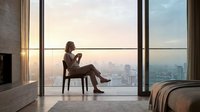 Young professional woman sitting by floor-to-ceiling windows reading book at sunrise in modern luxury apartment with city skyline view for lifestyle content.