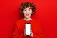 Surprised young woman with curly red hair showing blank smartphone screen against vibrant red background for advertising or app promotion.