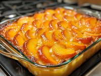 Fresh peach cobbler with caramelized fruit arranged in overlapping pattern, baked golden brown in glass baking dish, showing juicy syrup and crispy edges.