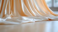 Elegant flowing silk fabric in cream and peach tones creates soft waves and folds against reflective surface, suitable for luxury fashion and wedding backgrounds.