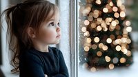 Young girl in navy sweater gazing thoughtfully through window with Christmas tree lights creating magical bokeh effect in background, capturing wonder of holiday season.