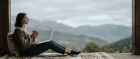 Young woman working remotely on laptop while enjoying morning coffee on wooden terrace with scenic mountain valley view in background.