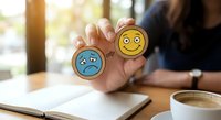 Professional woman holding wooden tokens with happy and sad emoji faces representing emotional choice and mental health awareness in workplace setting.