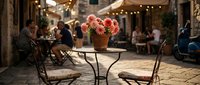 Outdoor café terrace with pink flowering plants in terracotta pot on metal table, European village street setting with warm string lights and blurred diners in background.