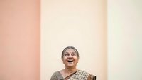 Woman with gray hair wearing a patterned top, smiling with joy and looking upward against a soft pastel background with pink and cream tones.